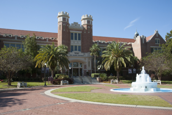 A red-brick university building with two towers, palm trees, and a circular fountain in front, set against a clear blue sky. Benches and brick pathways surround the fountain and entrance.