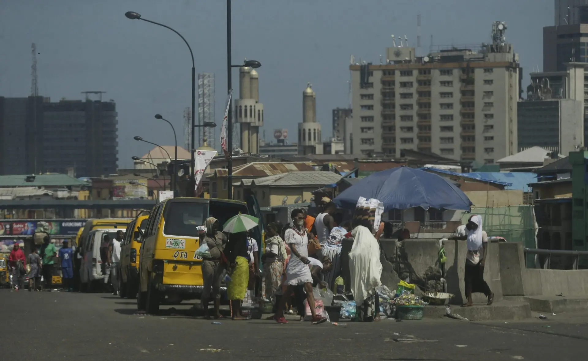 A busy street scene in an urban area with people walking, street vendors under umbrellas, yellow buses, and tall buildings in the background under a clear sky.