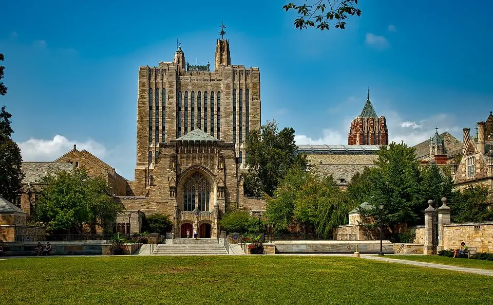 A large, historic stone building with a tall central tower, arched entrance, and surrounded by trees and lawns under a clear blue sky, seen from across a grassy open space.