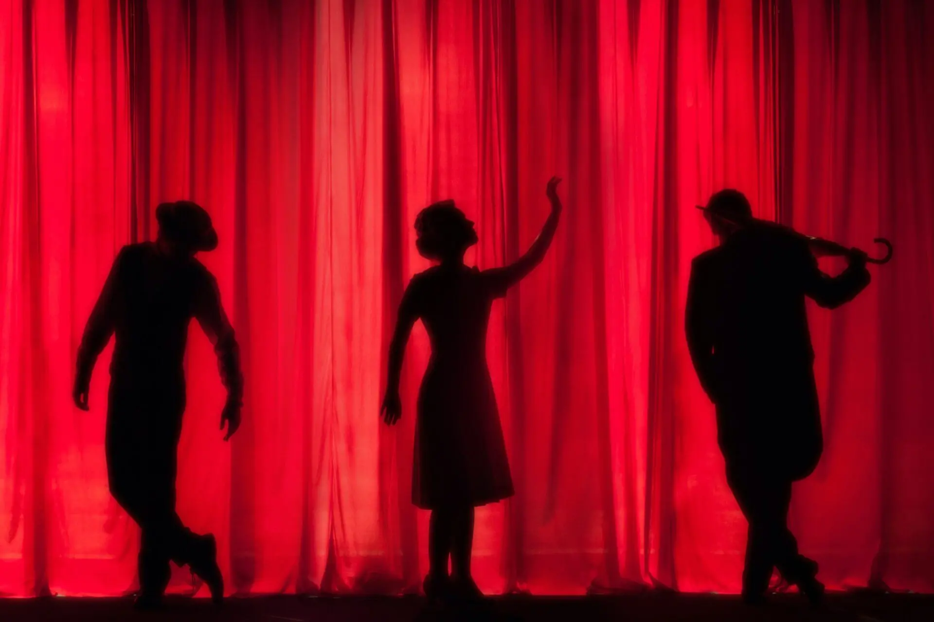 Three people stand in front of a bright red curtain, their silhouettes striking dramatic poses; one holds a hat, another raises an arm, and the third holds an umbrella.