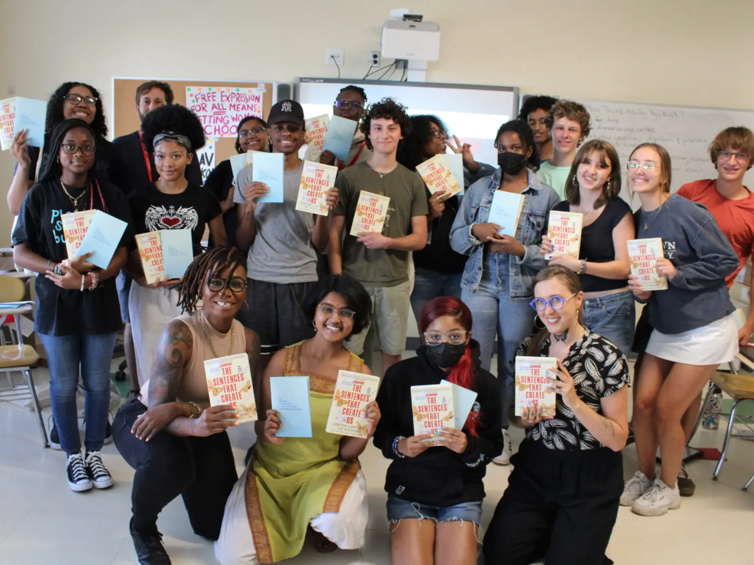 A diverse group of students and teachers pose in a classroom, smiling and holding copies of the book The Antiracist Writing Workshop. Some hold notebooks, and desks and a whiteboard are visible in the background.