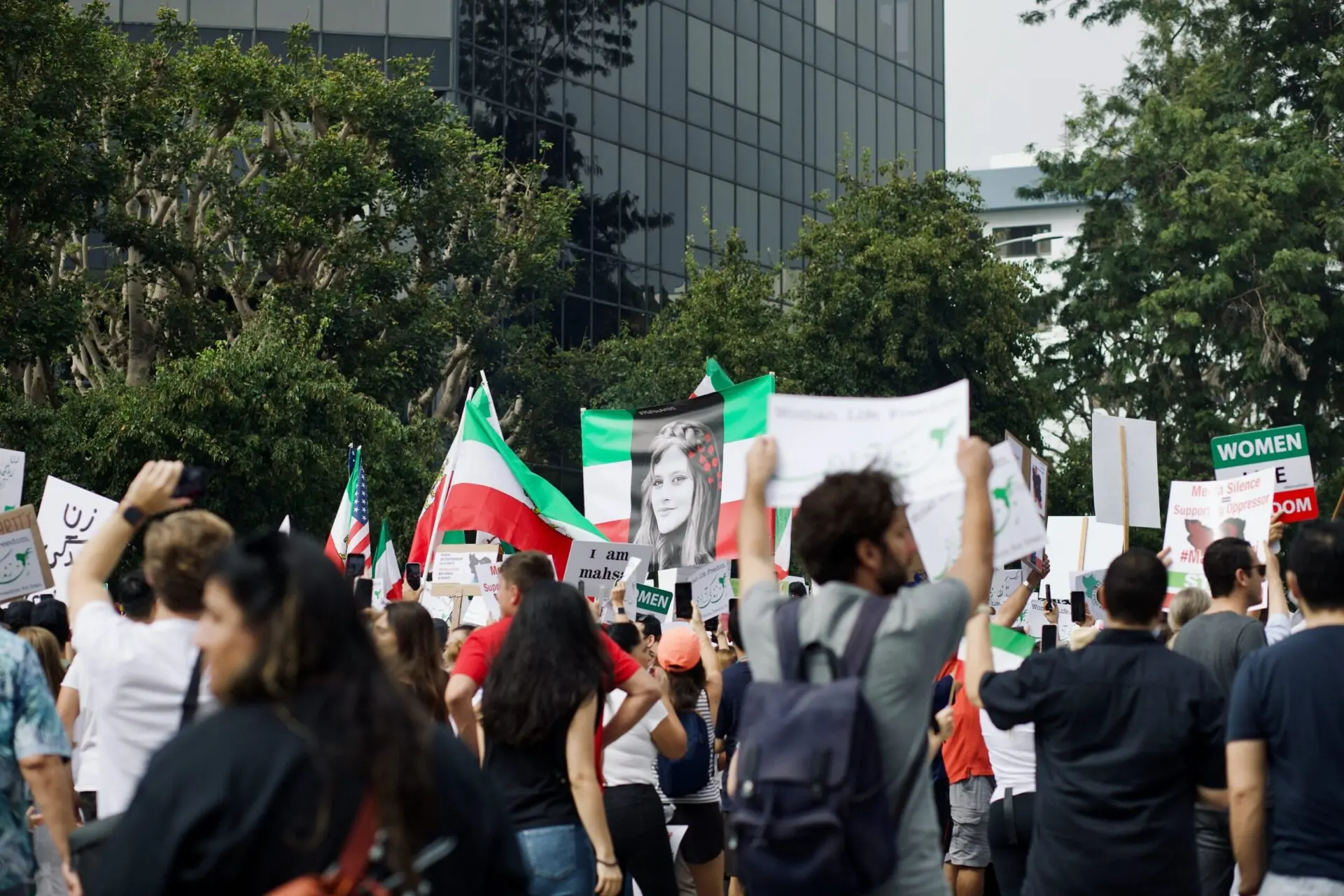 A large crowd of protesters holding signs and flags gathers outdoors; one prominent sign displays a black-and-white photo of a young woman. Tall buildings and trees are visible in the background.