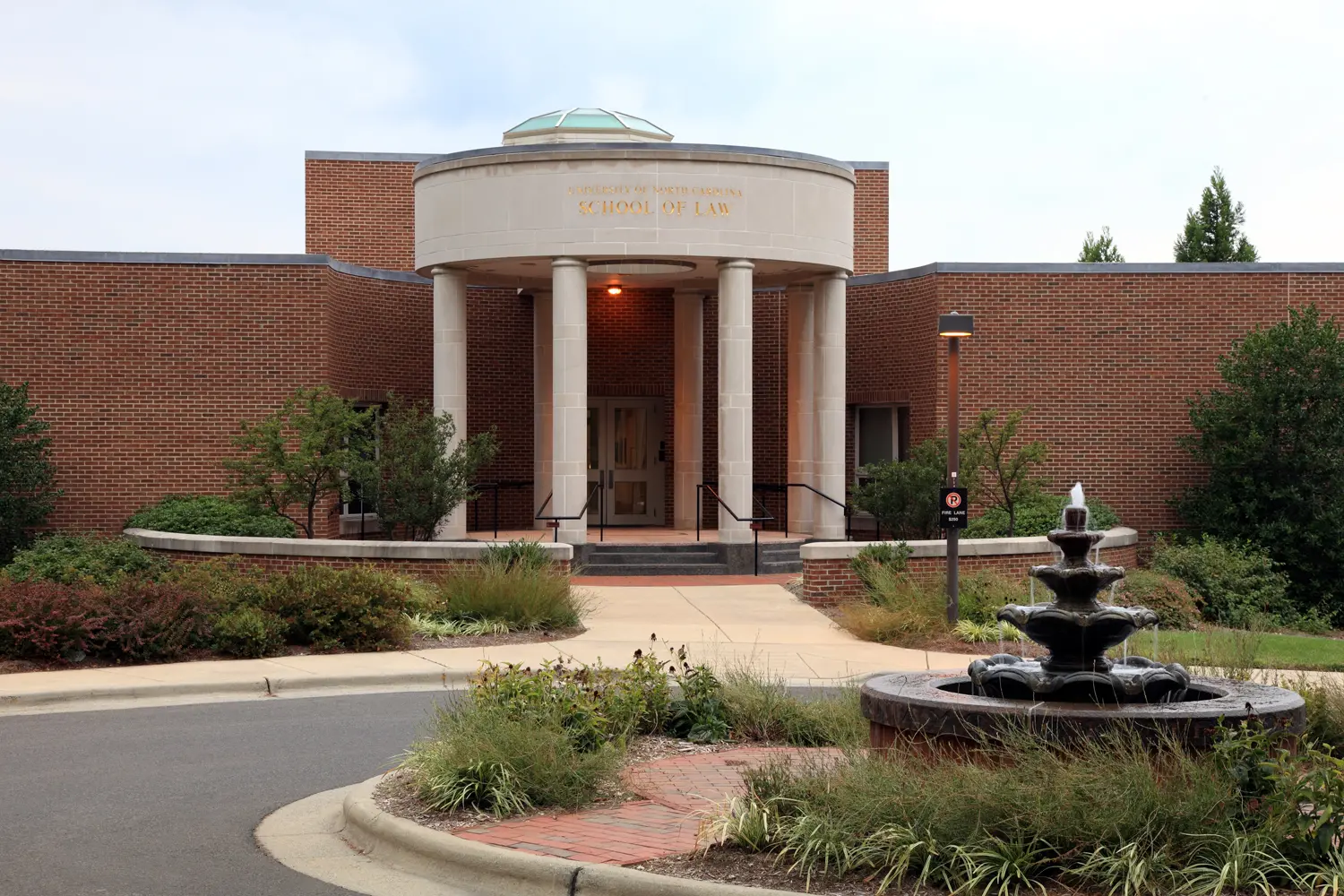 A brick building with a circular entrance supported by columns and a sign reading School of Law, surrounded by landscaping and a black tiered fountain in the foreground.