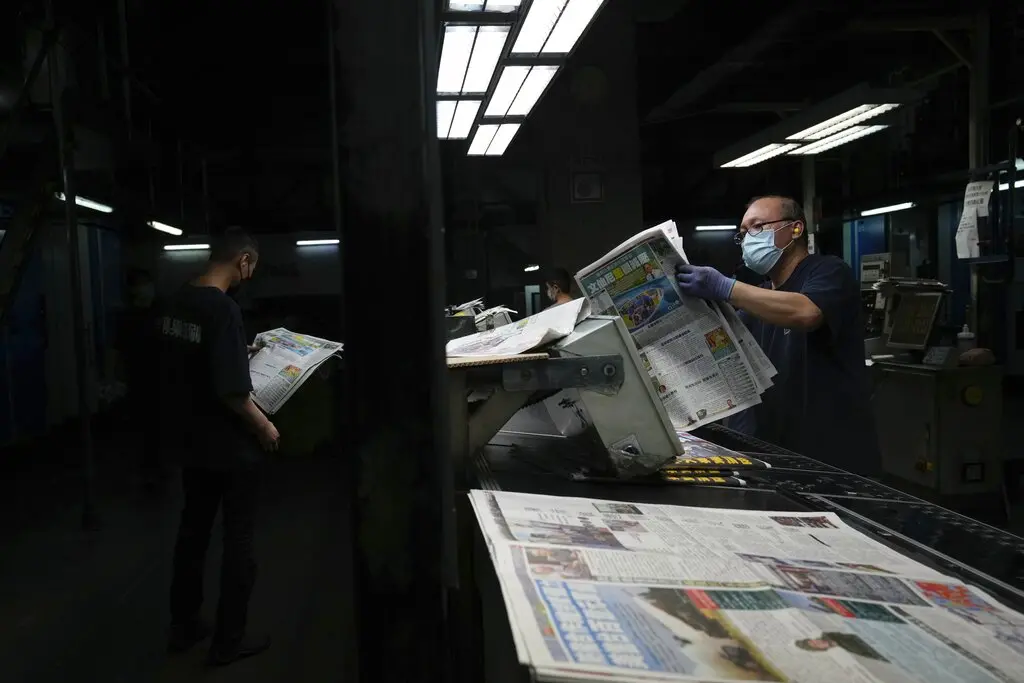 Two workers in a dimly lit printing facility inspect freshly printed newspapers. One person wears a mask and gloves, while stacks of newspapers move along a conveyor belt. Fluorescent lights illuminate the scene.