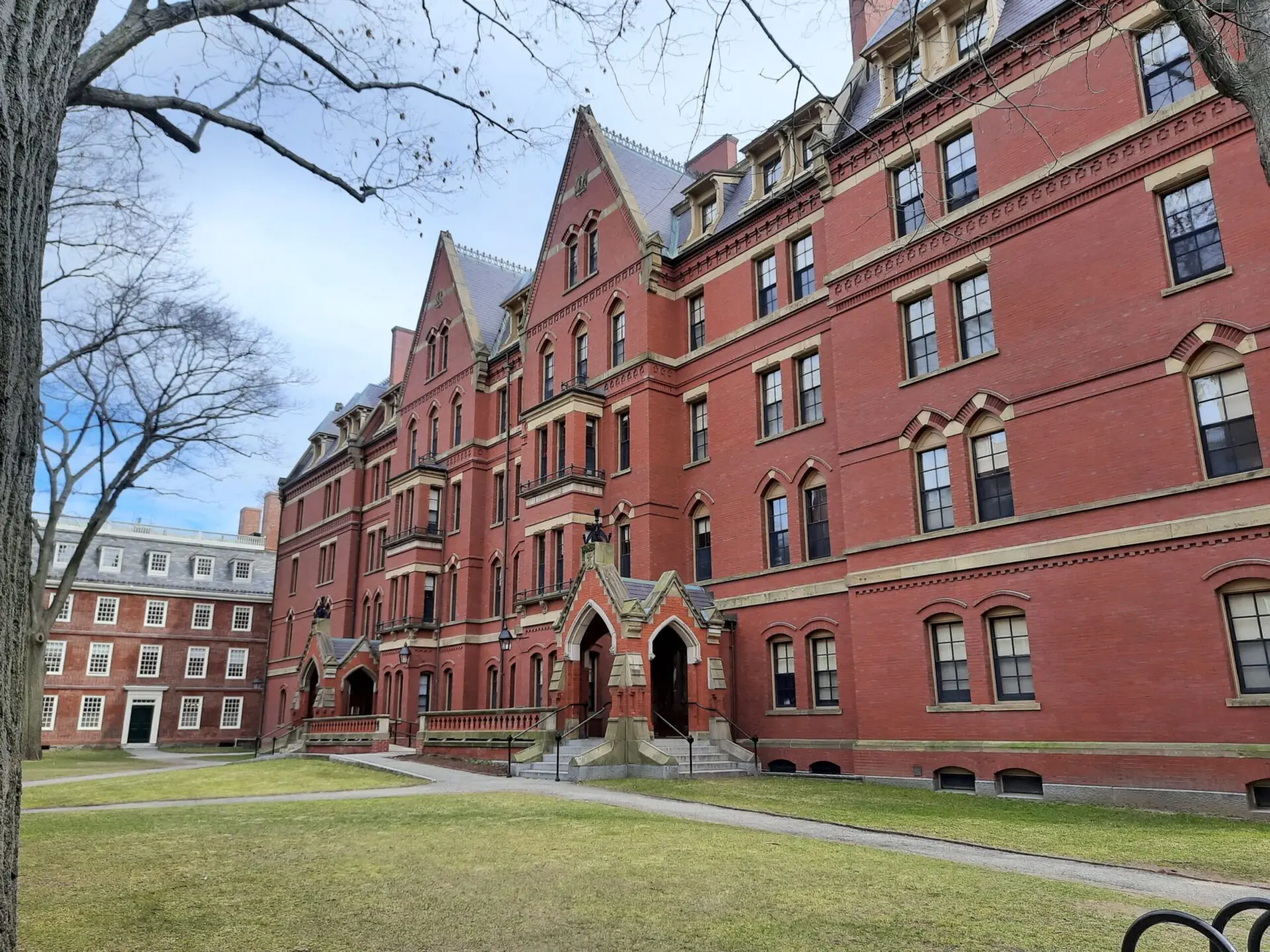 A large, red-brick university building with pointed rooftops and arched entrances sits beside a grassy courtyard on a clear day. Leafless trees frame the scene, and a stone path leads to the building.