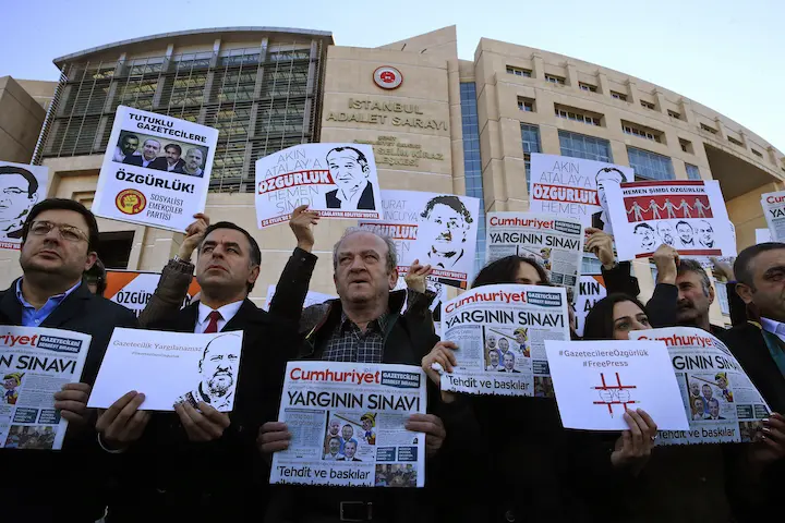 A group of people protest outside a large courthouse, holding newspapers, posters with faces, and signs calling for press freedom and the release of jailed journalists.