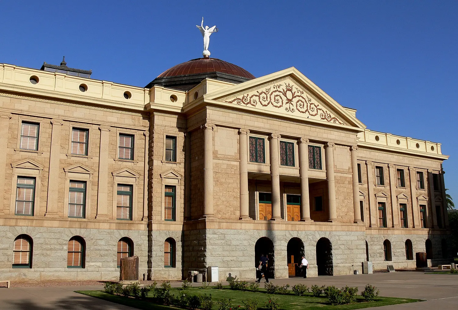 A grand, historic building with columns, arched doorways, and a domed roof topped by a white statue, set against a clear blue sky. A few people walk near the entrance. Green lawn and shrubs are in front.