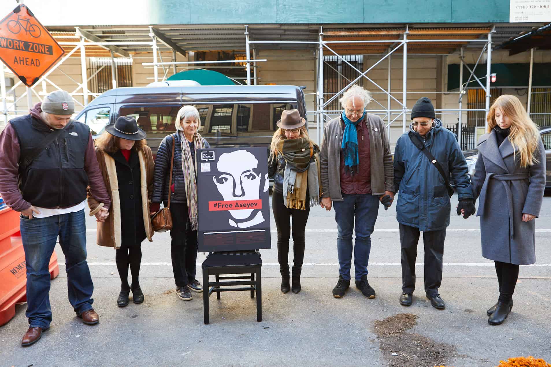 Seven people stand in a line on a city street, holding hands and bowing their heads in front of a sign with a face and the words #Free Assange. Scaffolding and a van are visible in the background.