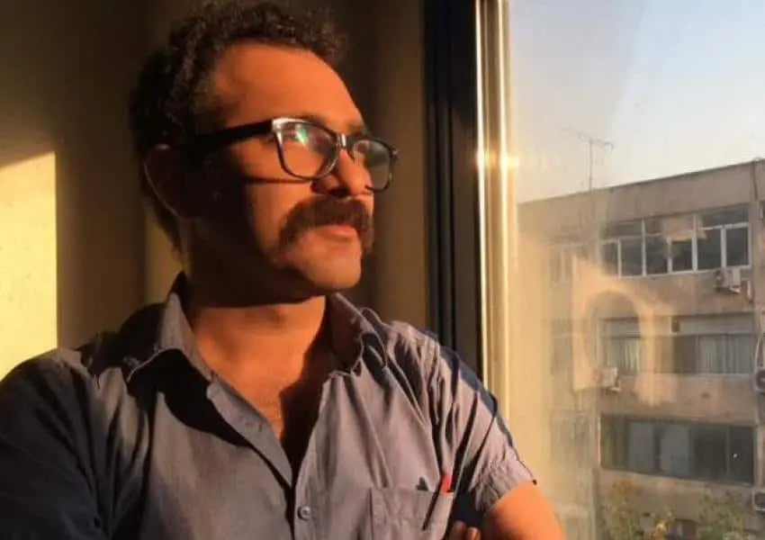 A man with glasses and a mustache stands by a window, looking outside with arms crossed. Sunlight casts a warm glow on his face and shirt, and a building is visible through the window.