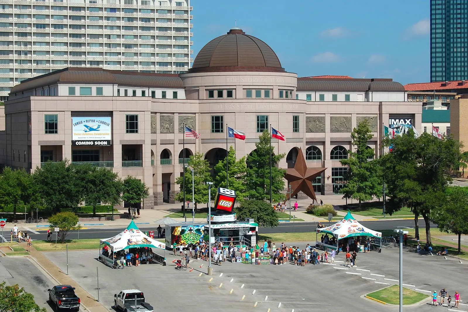 A large museum building with a dome and Texas flags, surrounded by trees. People gather at colorful tents in the parking lot below. A giant bronze star sculpture stands at the entrance.