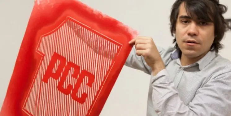A person with dark hair holds up a red print featuring the outline of a T-shirt with the letters PCC inside, created with vertical stripes.