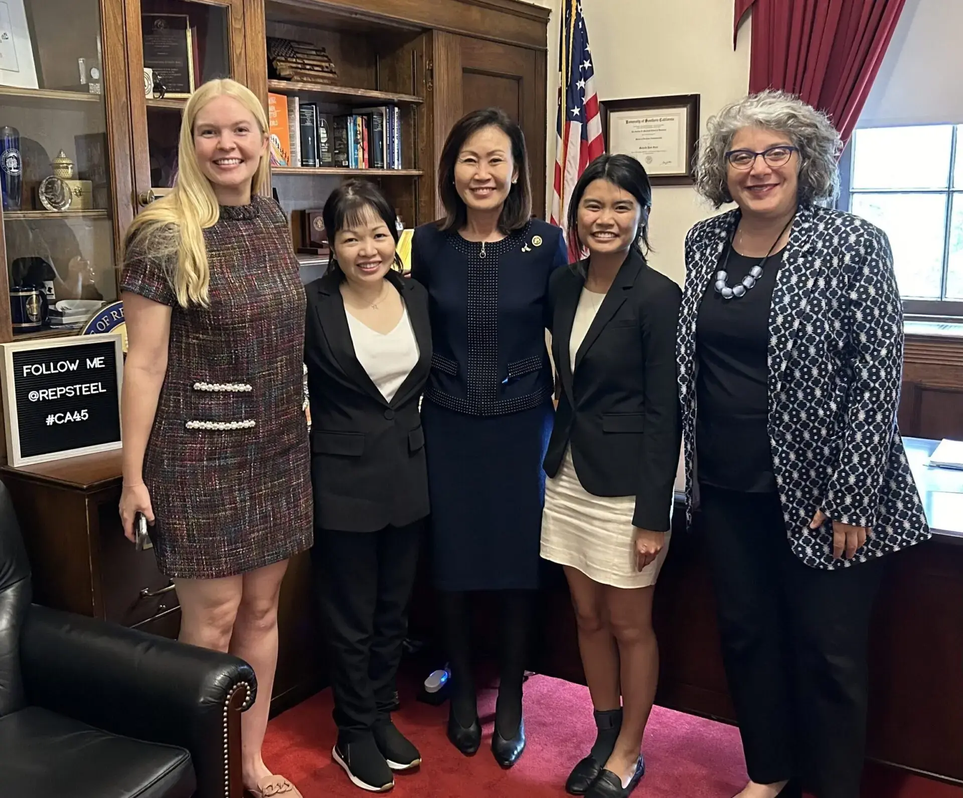 Five women stand together and smile in an office with bookshelves, an American flag, and a “Follow Me @RepSteel #CA45” sign, as PEN America news is discussed. They are all dressed in professional attire.