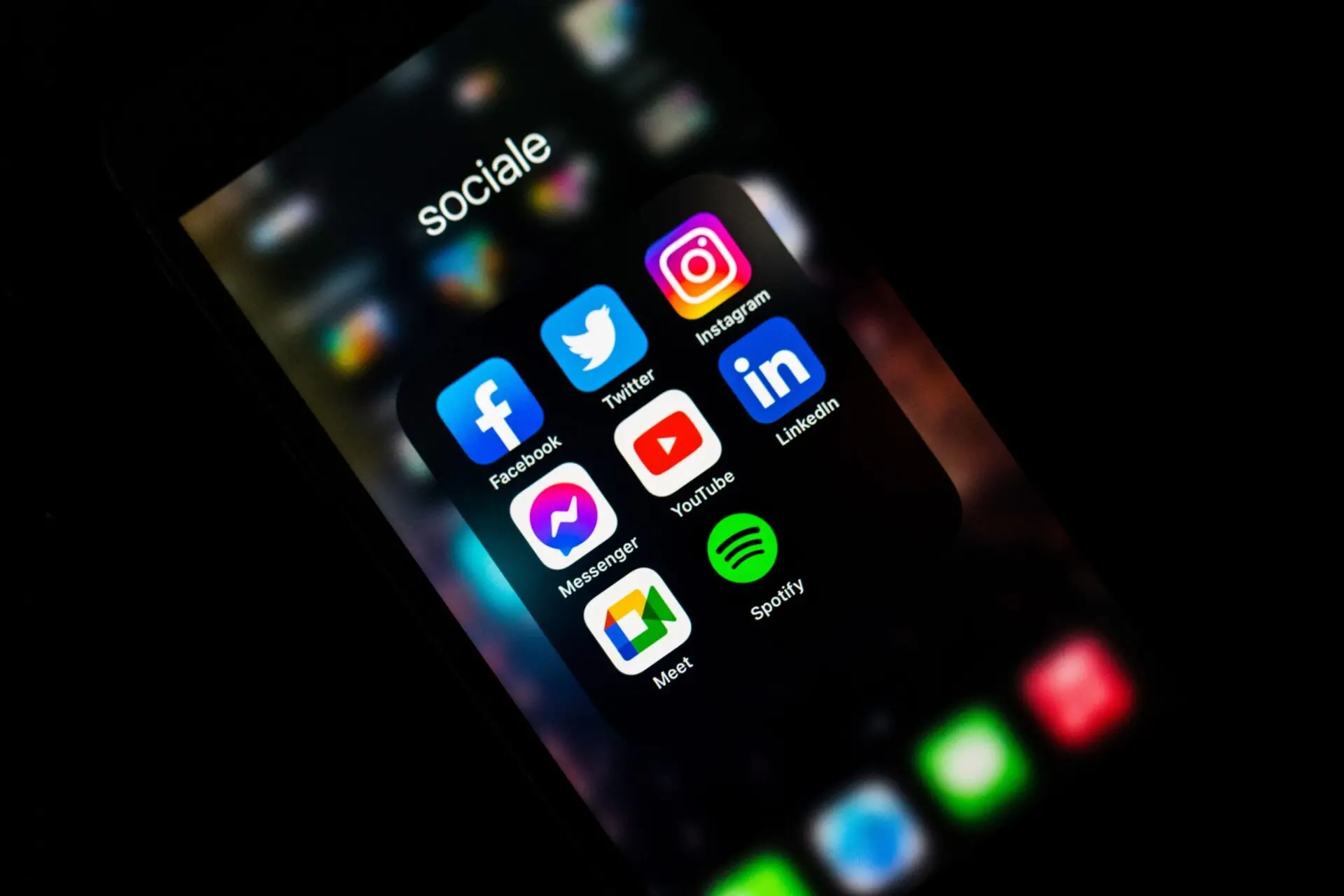 A smartphone screen displays a folder labeled sociale containing app icons for Facebook, Twitter, Instagram, Messenger, YouTube, LinkedIn, Meet, and Spotify. The background is blurred and dark.