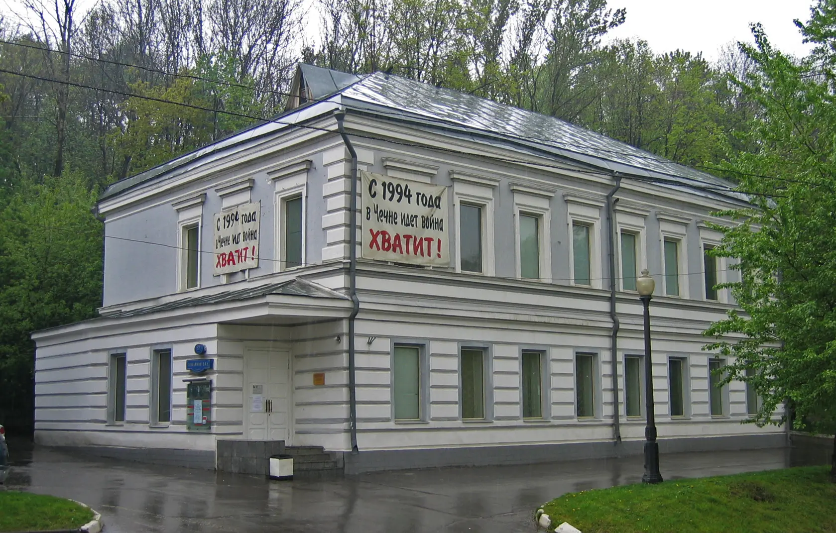 A white, two-story building with banners in Russian on the walls, set on a rainy day with wet pavement and trees in the background. The banners say, Since 1994 in Chechnya, there is war! Enough!.