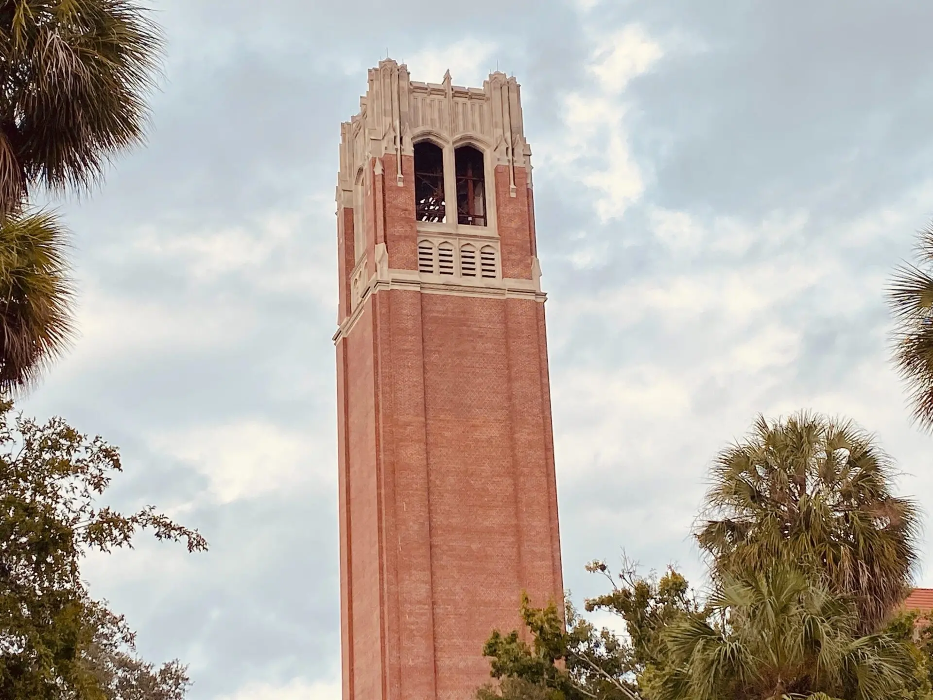 A tall red-brick clock tower with white details rises above green trees against a cloudy sky. The tower has arched windows near the top and decorative stonework.