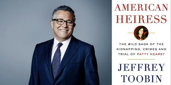 A man in a suit and glasses smiles next to the book cover of American Heiress: The Wild Saga of the Kidnapping, Crimes and Trial of Patty Hearst by Jeffrey Toobin.
