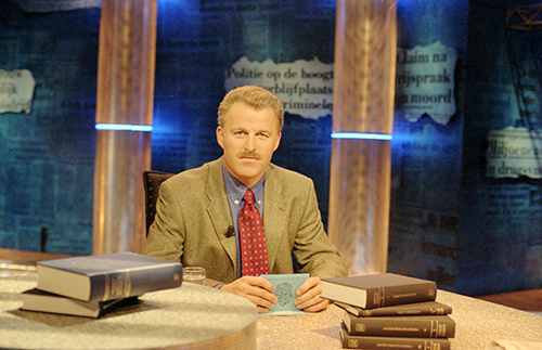 A man in a suit and red tie sits at a round table with several large books stacked in front of him. The background is blue with text and columns, suggesting a formal talk show or news set.
