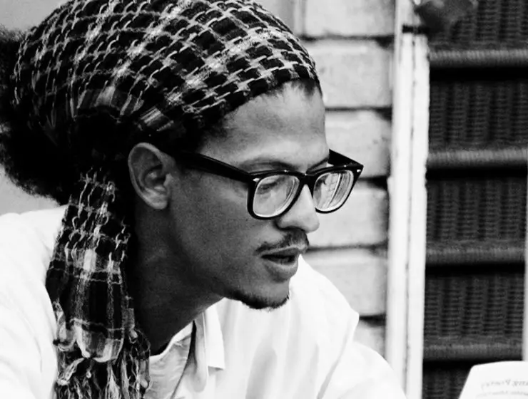 Black and white photo of a person wearing glasses and a patterned headscarf, looking down thoughtfully. The background features a brick wall and part of a window with bars.