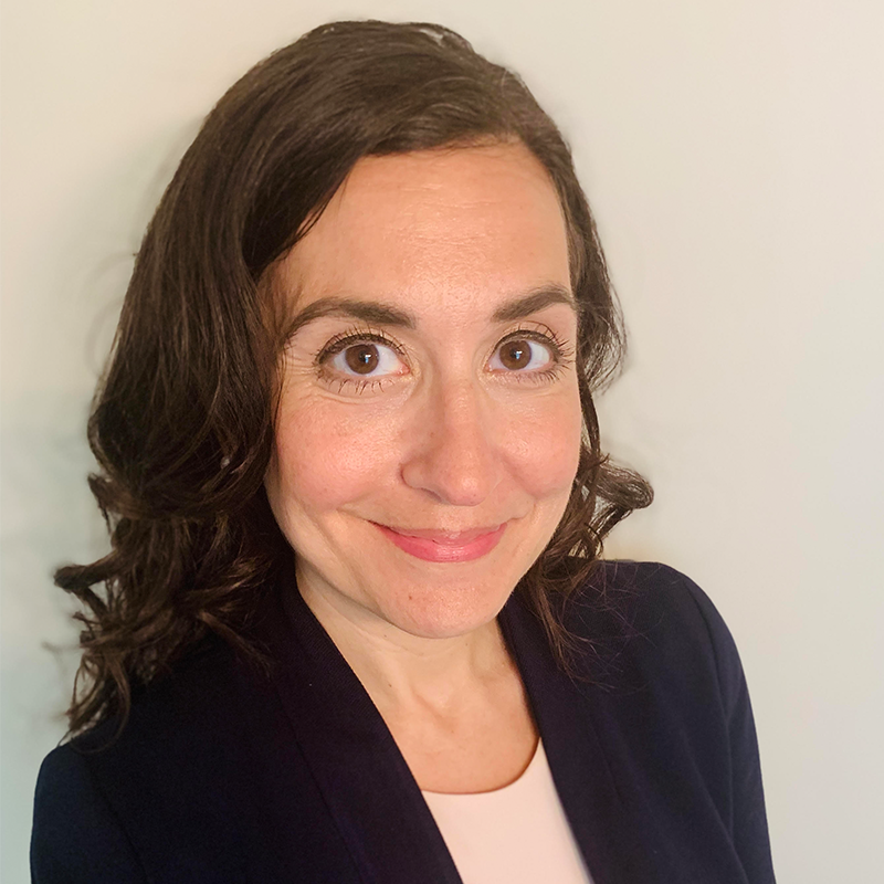 A woman with wavy brown hair smiles at the camera. She is wearing a navy blazer over a light-colored top, posed in front of a plain, light background at the DC Free Expression Advocacy Institute PEN event.