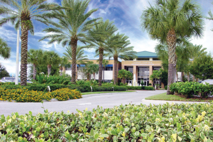A building with white columns and a green roof is surrounded by palm trees, manicured bushes, and a circular driveway under a blue sky with scattered clouds.