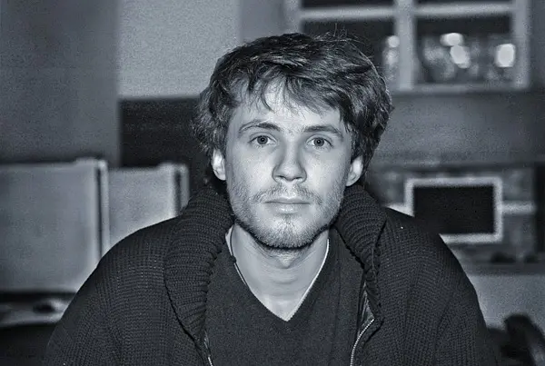 A young man with tousled hair and a light beard looks directly at the camera. He is wearing a dark sweater and is seated indoors, with blurred office equipment in the background. The photo is in black and white.