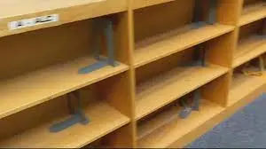 Empty wooden bookshelves with metal bookends, situated in a library or classroom setting. The shelves have labels on them but no books are visible. The floor appears to be carpeted in blue.