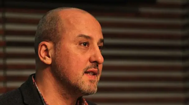 A middle-aged man with a shaved head and short beard, wearing a checked shirt and dark jacket, speaking in front of a background with horizontal lines.