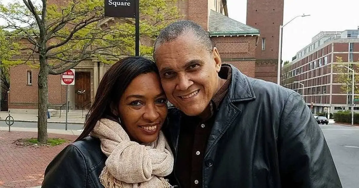 A smiling couple wearing dark jackets poses together outdoors near a brick sidewalk, with a church and street signs visible in the background. The woman has a beige scarf and the man has his head gently touching hers.
