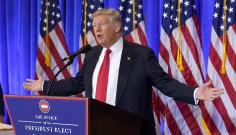 A man in a suit and red tie stands at a podium labeled The Office of the President Elect, speaking with arms outstretched. Multiple American flags are displayed in the background.