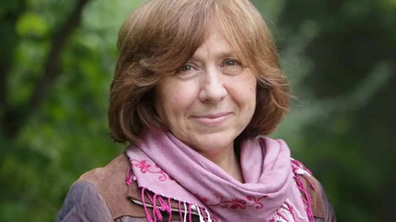 A middle-aged woman with light brown hair smiles gently at the camera, wearing a mauve scarf embroidered with flowers and a brown jacket, standing outdoors with greenery in the background.