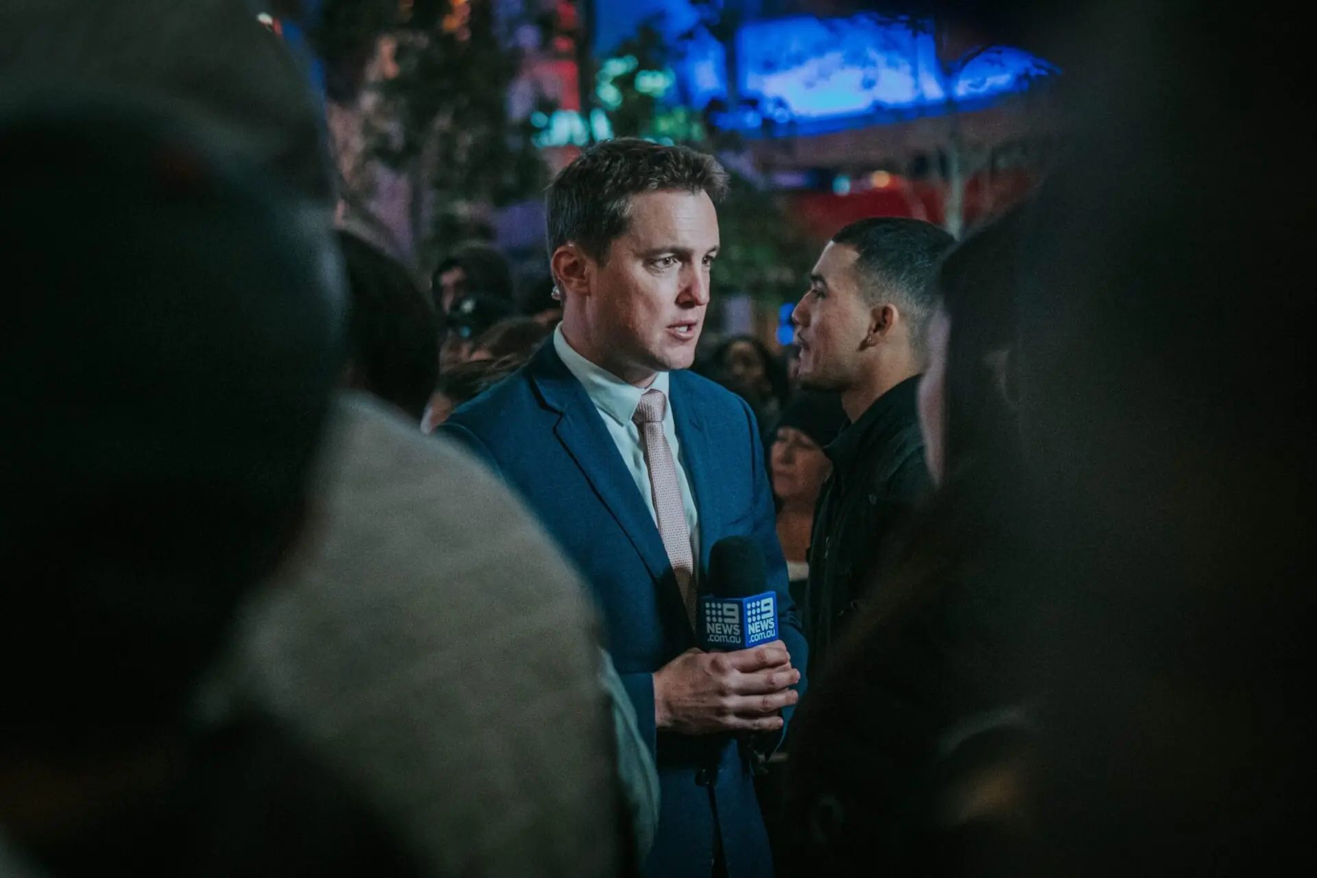 A male reporter in a blue suit holds a microphone labeled NEWS while speaking to the camera, surrounded by a crowd of people at night with colorful lights in the background.