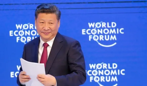A man in a dark suit and red tie holds papers and smiles while standing in front of a blue backdrop with the World Economic Forum logo.