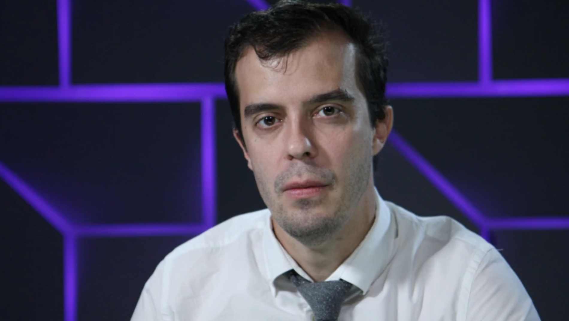 A man with short dark hair and a light complexion, wearing a white shirt and gray tie, looks at the camera against a background with purple geometric patterns.