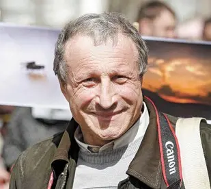 An older man with gray hair smiles at the camera, wearing a brown jacket and a Canon camera strap around his neck. A blurred outdoor background and photos can be seen behind him.