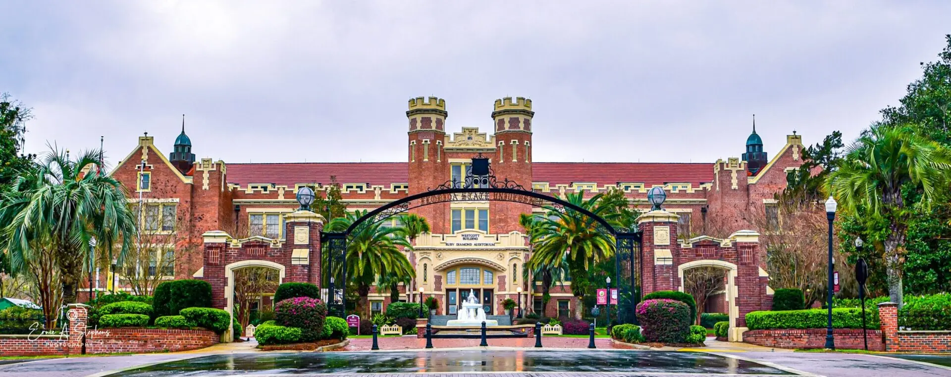 A historic, red-brick university building with two towers and an arched iron gate in front, surrounded by palm trees and lush greenery on a rainy day.