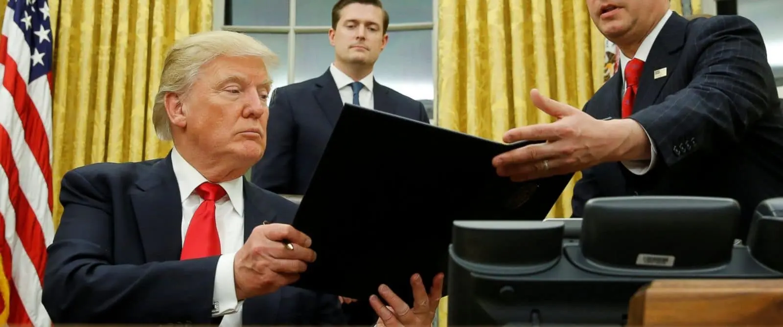 Donald Trump sits at a desk in the Oval Office, holding a large black folder as a man hands it to him. Another man stands behind them, and yellow curtains are visible in the background.