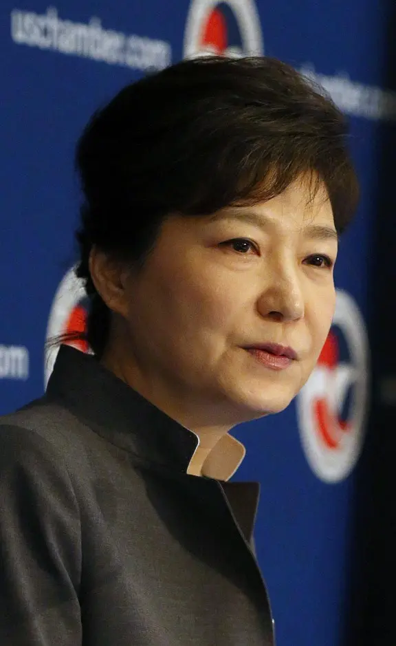 A woman with short dark hair wearing a dark jacket speaks in front of a blue backdrop with partial white and red logos and text.