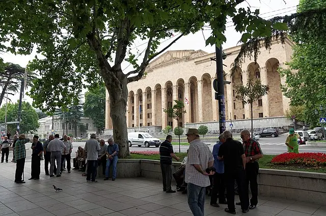 A group of people stands on a tree-lined sidewalk near a street, with a large historic building featuring tall columns and arched windows in the background. Cars are parked along the street and flower beds are visible.