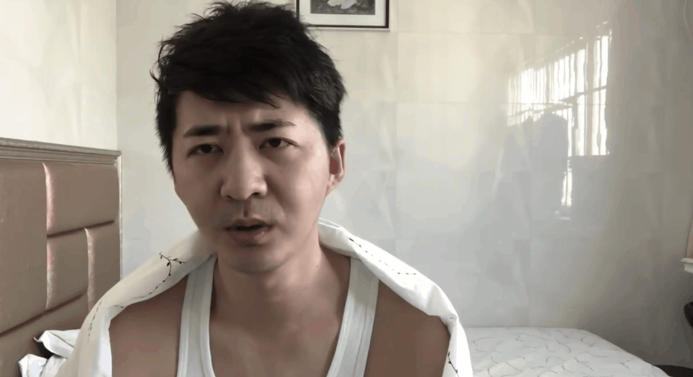 A man with short dark hair sits on a bed, wearing a white tank top and draped in a blanket, looking directly at the camera with a serious expression. Sunlight enters through a window behind him.