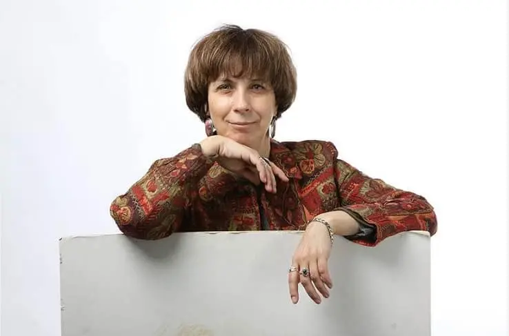 A woman with short brown hair, wearing a patterned red and gold jacket, smiles gently while resting her chin on her hand and leaning on a white surface against a plain white background.