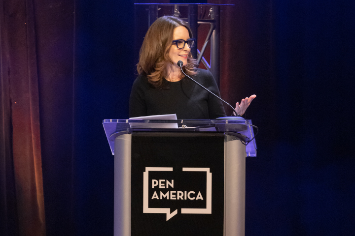 A woman with brown hair and glasses speaks at a podium with a microphone. The podium displays the PEN America logo. The background is dark with stage lighting.