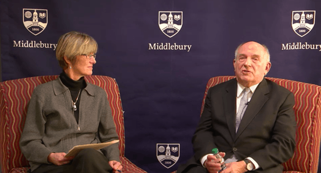 A woman and a man sit in armchairs, engaged in conversation, with a blue Middlebury College banner in the background. The man holds a water bottle, and the woman has a folder or papers in her lap.