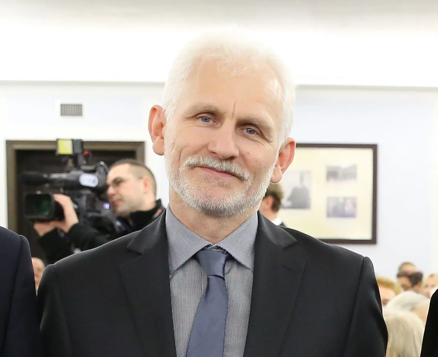 A man with short white hair and a beard, wearing a suit, light gray shirt, and tie, stands indoors and smiles slightly. People and a cameraman are visible in the blurred background.