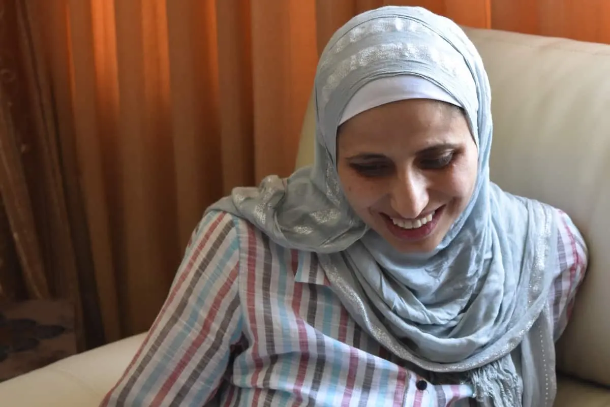 A woman wearing a light blue hijab and a striped shirt sits on a white couch, smiling and looking down. Warm, brown curtains are in the background.