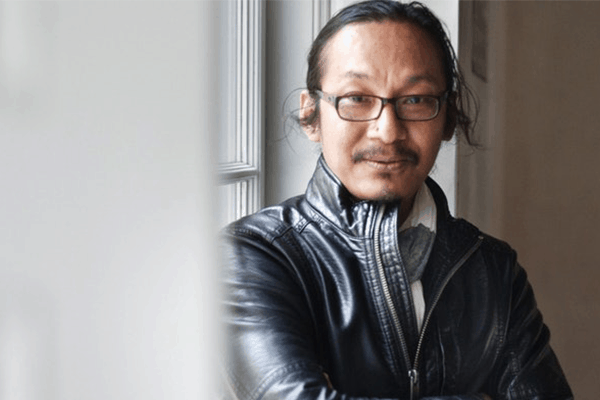 A man with glasses and long hair tied back is wearing a black leather jacket and scarf, standing indoors next to a window, looking at the camera and gently smiling.