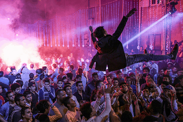 A lively crowd at a night event tosses a person into the air under colorful lights and smoke, with many people cheering and reaching up in celebration.