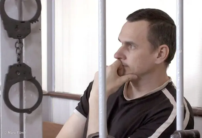 A man sits pensively behind bars in Russia, resting his chin on his hand. Open handcuffs hang on the metal bars beside him. The lighting is bright, with a white wall and window in the background.
