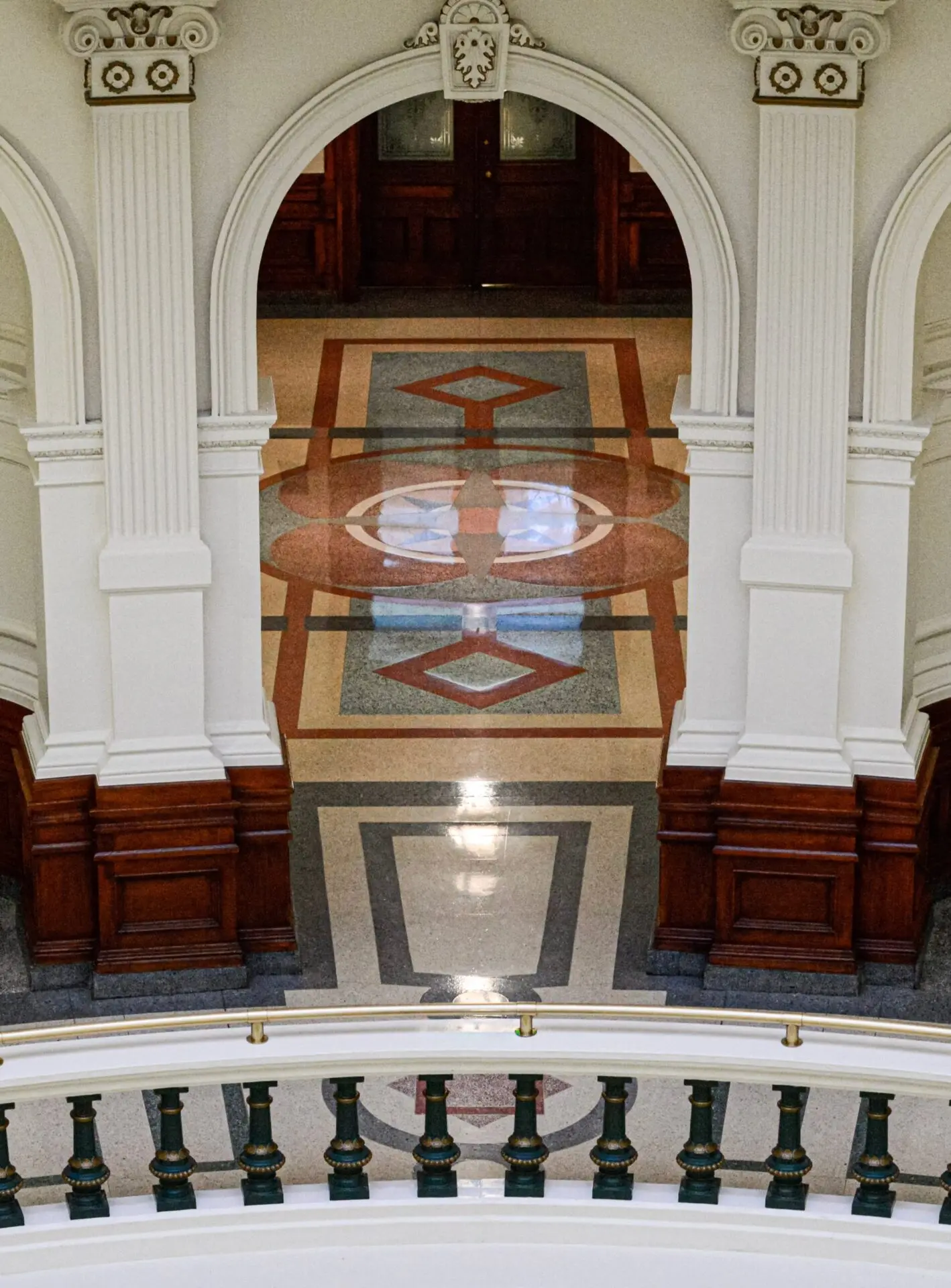 A view of an ornate interior with white columns, an arched entry, decorative moldings, and a geometric patterned floor in shades of brown, red, and gray. A green and white banister is visible in the foreground.