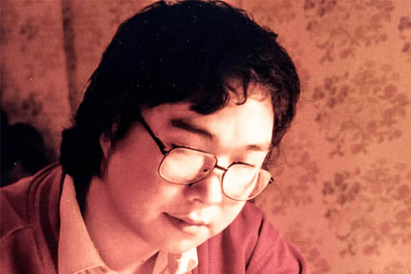 A person with medium-length dark hair and glasses looks down, appearing thoughtful. They are wearing a light-colored shirt and a reddish sweater. The background is softly lit with a floral patterned wallpaper.