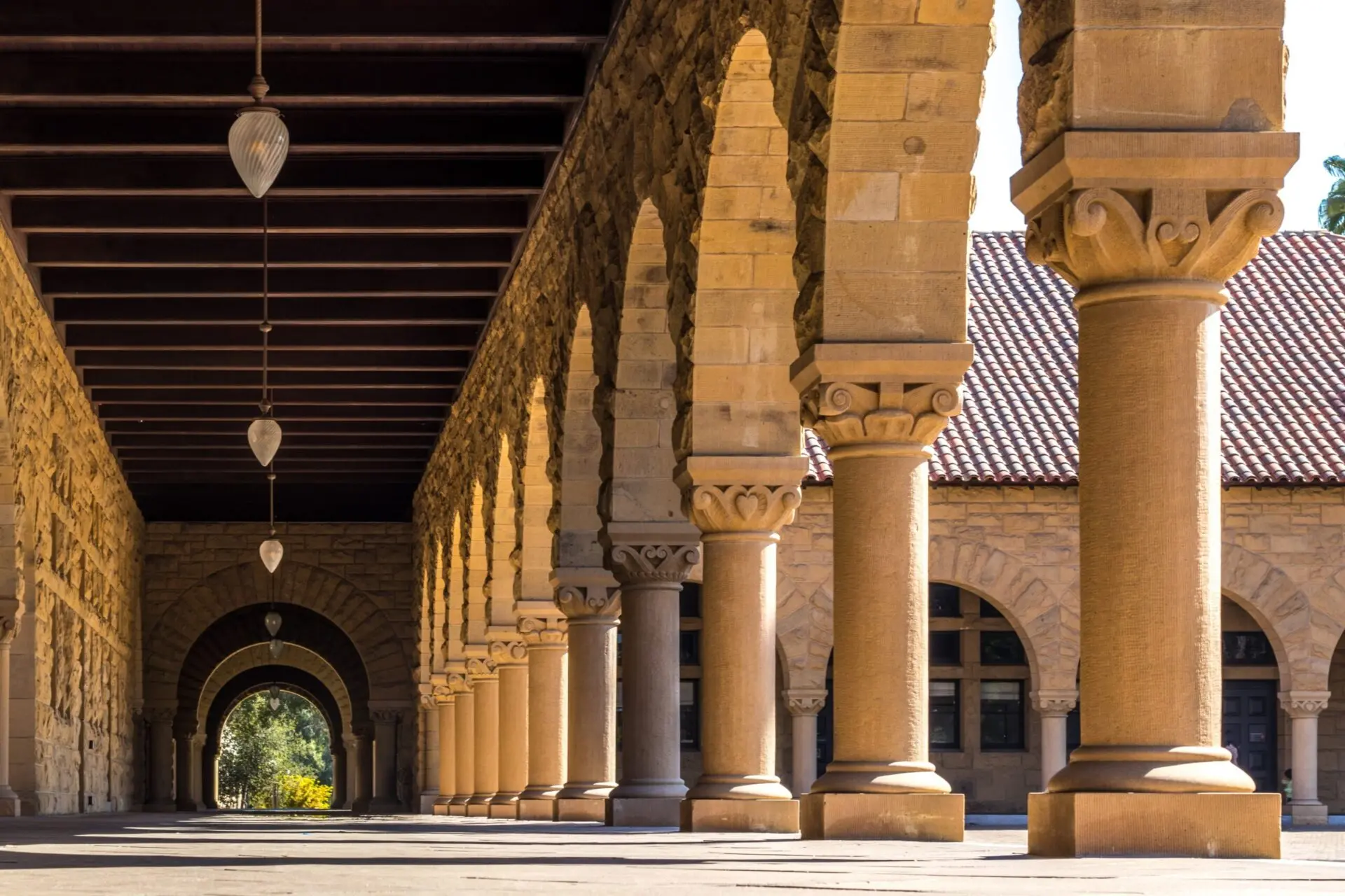A view of stone arches and columns in an outdoor corridor at Stanford University, with hanging lanterns and sunlight casting shadows along the walkway.
